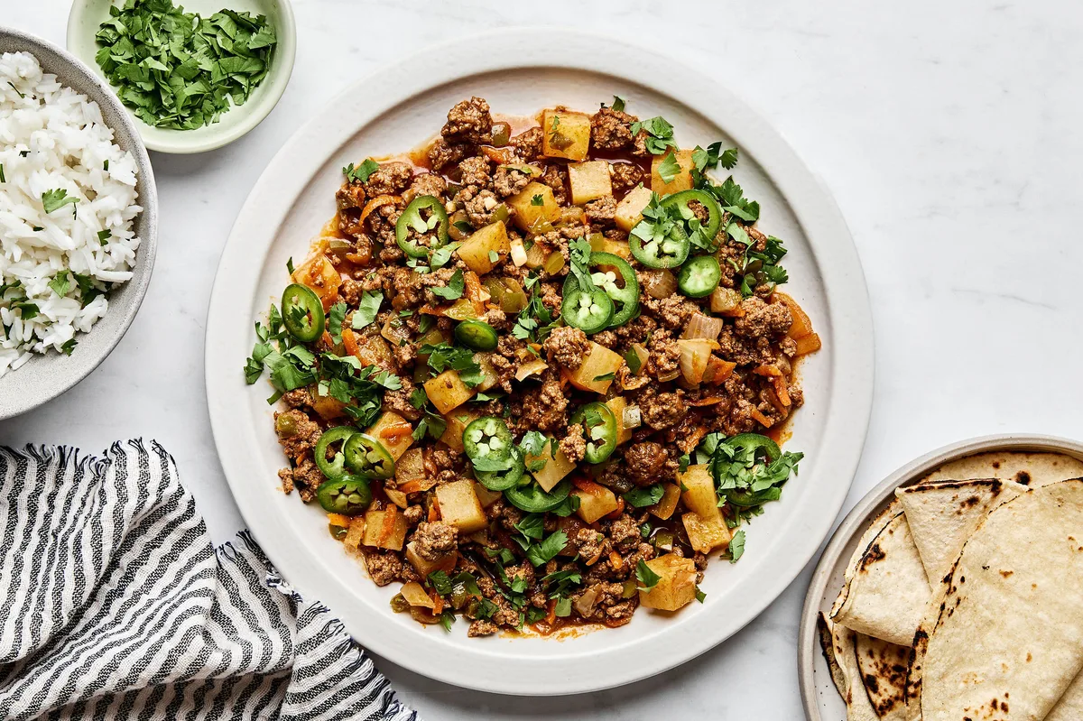 A bowl of picadillo topped with fresh cilantro and jalapeños, served with rice and a warm tortillas on the side