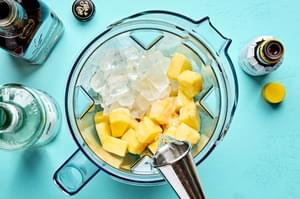pineapple, ice, cream of coconut, pineapple juice, lime juice, and light rum and bitters being added to a blender