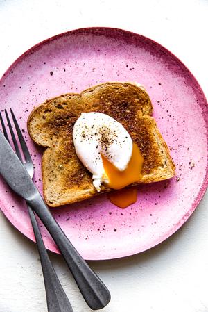 Pink plate with a piece of toast and a poached egg with salt and pepper a fork and knife