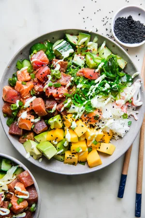 A poke bowl featuring a base of seasoned rice, topped with tuna salmon, mango, cucumber salad, avocado & citrus wasabi sauce