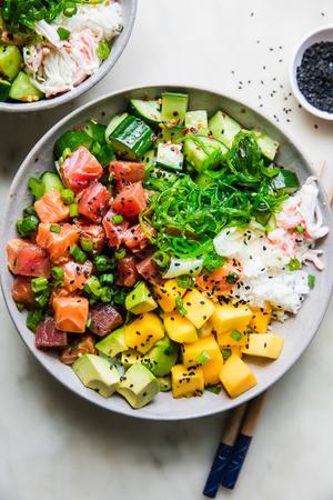 A poke bowl featuring a base of seasoned rice, topped with tuna salmon, sweet mango, crisp cucumber salad, avocado, & seaweed