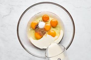 6 raw eggs, cream, salt, pepper, and nutmeg being whisked together in a bowl