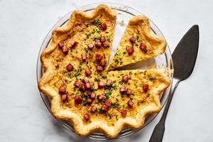 Quiche Lorraine in a pie pan made with eggs, cream, ham, brown sugar, nutmeg and Gruyère topped with chives being sliced