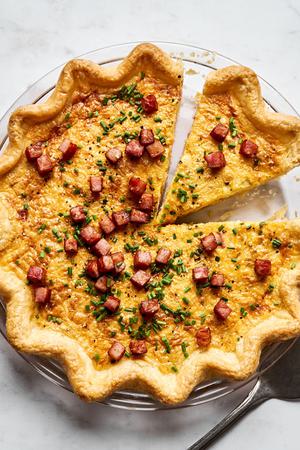Quiche Lorraine in a pie pan made with eggs, cream, ham, brown sugar, nutmeg and Gruyère topped with chives being sliced