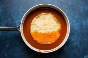 a corn tortilla being dipped in red enchilada sauce in a frying pan