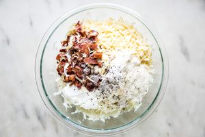 grated potatoes and onion in a glass bowl with bacon, gruyere, parmesan, butter, salt, pepper, and nutmeg
