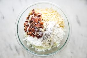 grated potatoes and onion in a glass bowl with bacon, gruyere, parmesan, butter, salt, pepper, and nutmeg