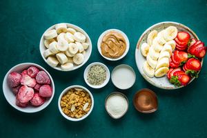 bowls of strawberries, bananas, granola, peanut butter, milk coconut oil, protein powder on the counter for smoothie bowl