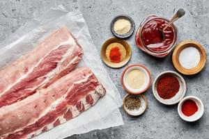 Ingredients laid out for making oven-baked ribs, including a rack of raw pork ribs, a bowl of dry rub spices & barbecue sauce