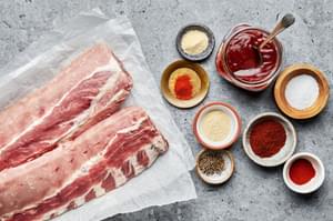 Ingredients laid out for making oven-baked ribs, including a rack of raw pork ribs, a bowl of dry rub spices & barbecue sauce