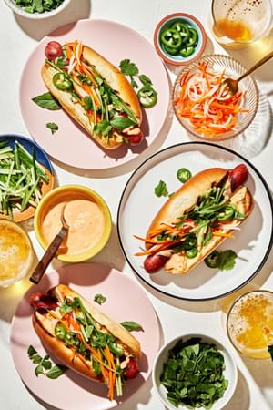 Banh Mi Hot Dogs surrounded by fixings, Sriracha mayo, pickled veggies, cilantro, basil, cucumbers and jalapeños