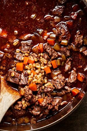 a pot of homemade beef & barley soup being stirred with a spoon made with tomato paste, soy sauce, vegetables and spices