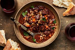 a bowl of homemade beef & barley soup with a spoon surrounded by crusty bread for serving