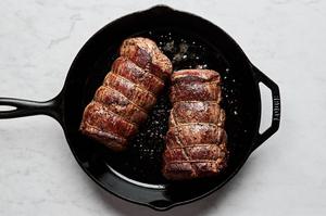 2 Beef tenderloins tied with twine and seasoned with salt and pepper being seared in a skillet with oil