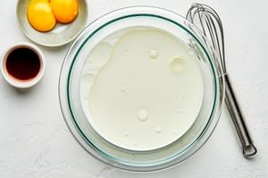 milk and oil in a boil next to small bowls of egg yolks and vanilla with a whisk sitting beside them on the counter