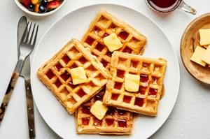 4 homemade Belgian waffles on a plate with a pad of butter and syrup on each next to a knife and fork