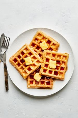 4 homemade Belgian waffles on a plate with a pad of butter and syrup on each next to a knife and fork