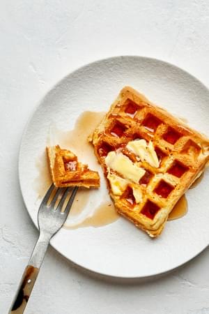 A homemade Belgian waffle on a plate with a pad of butter and syrup on top with one bite cut off with a fork