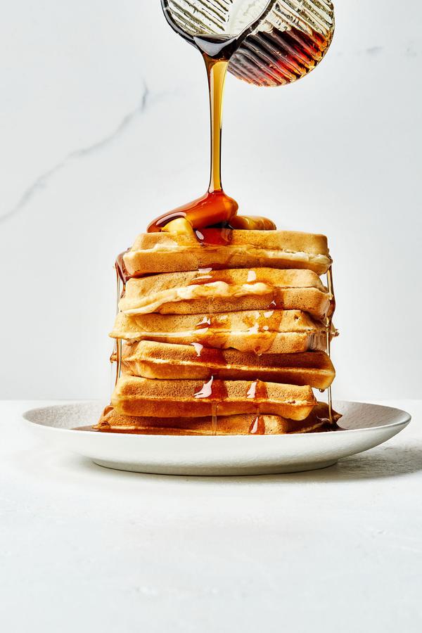 a stack of homemade Belgian waffles on a plate topped with butter and having warm syrup poured over them