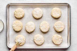 homemade raw biscuits on a parchment lined baking sheet being brushed with buttermilk to turn them golden while baking