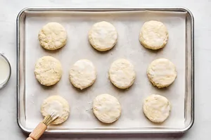 homemade raw biscuits on a parchment lined baking sheet being brushed with buttermilk to turn them golden while baking