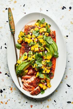 blackened salmon topped with homemade mango salsa and avocado on a serving plate with a serving fork
