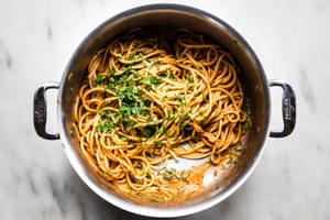 cooked bucatini noodles being tossed with spicy roasted red paper sauce and fresh parsley in a stock pot
