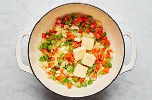 butter being added into a stock pot with chopped onion, carrots, celery and garlic that have been cooked in olive oil