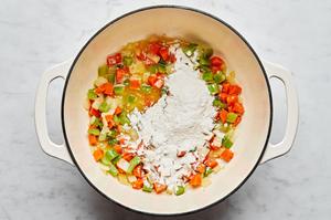 flour being added into a stock pot with chopped onion, carrots, celery, garlic, olive oil and butter