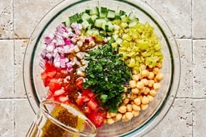 Homemade Italian dressing being poured on top of chickpea salad with cucumber, tomatoes, red onion, parsley and peperoncinis