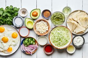ingredients in bowls for chilaquiles including fried eggs, corn tortillas, shredded cabbage, salsa verde and sour cream