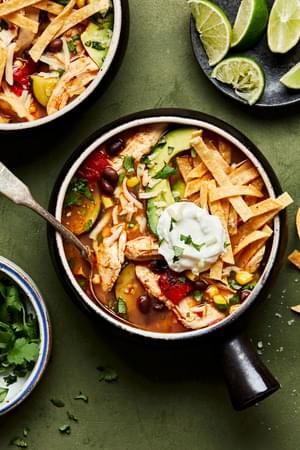 a bowl of homemade chipotle chicken soup topped with shredded cheese, tortilla chips, lime and avocado