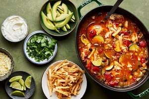a pot of chipotle chicken soup next to bowls of garnishes: sour cream, cilantro, avocado, tortilla chips, cheese and limes