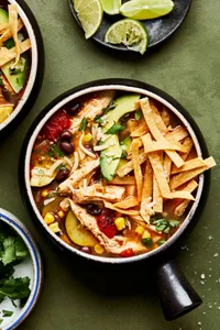 a bowl of homemade chipotle chicken soup topped with shredded cheese, tortilla chips, lime and avocado