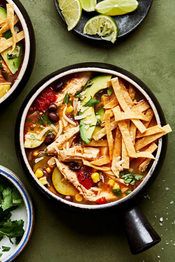 a bowl of homemade chipotle chicken soup topped with shredded cheese, tortilla chips, lime and avocado