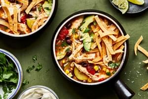 a bowl of homemade chipotle chicken soup topped with shredded cheese, tortilla chips, lime and avocado