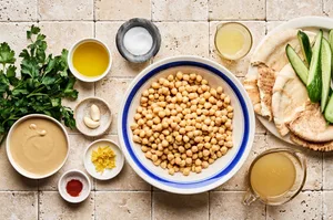 Chickpeas, tahini, olive oil, lemon juice, garlic, salt, cucumber, pita, parsley, paprika, & za'atar laid out to make hummus