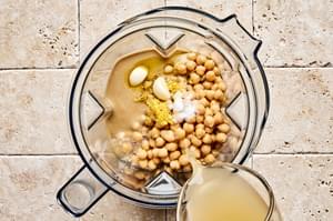 Chickpeas, tahini, olive oil, lemon juice, garlic, and salt being added to a blender to make hummus.
