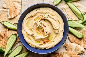 A bowl of homemade hummus drizzled with olive oil, topped with parsley, paprika, and za’atar, served with pita and cucumber.