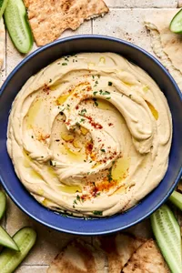 A bowl of homemade hummus drizzled with olive oil, topped with parsley, paprika, and za’atar, served with pita and cucumber.