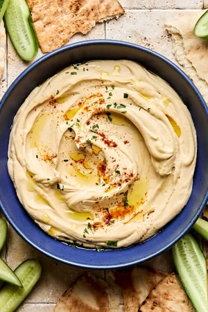 A bowl of homemade hummus drizzled with olive oil, topped with parsley, paprika, and za’atar, served with pita and cucumber.