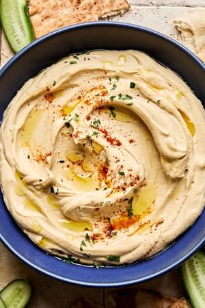 A bowl of homemade hummus drizzled with olive oil, topped with parsley, paprika, and za’atar, served with pita and cucumber.