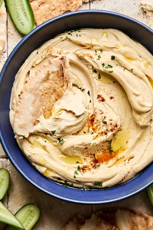 A bowl of homemade hummus drizzled with olive oil, topped with parsley, paprika, and za’atar, served with pita and cucumber.