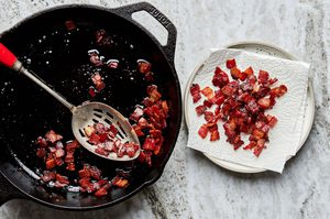 crispy bacon pieces cooking in a skillet being scooped out with a spoon onto a paper towel lined plate