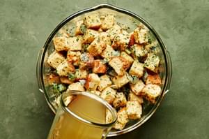 chicken stock being poured over cornbread, parsley and cooked onion and Celery in a large mixing bowl