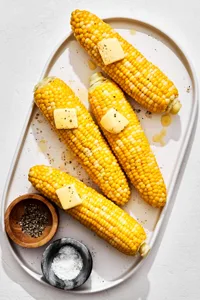 4 homemade corn on the cob on a platter topped with butter beside small bowls of salt and pepper for serving