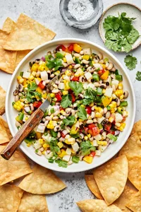 homemade cowboy caviar in a serving bowl with a spoon surrounded by tortilla chips and bowls of salt and cilantro