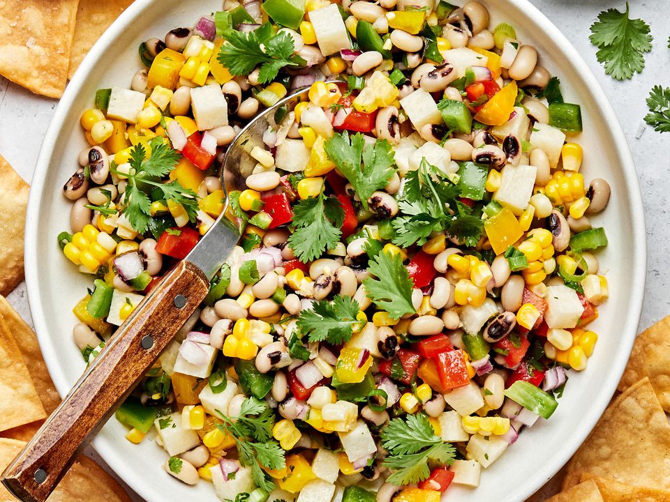 homemade cowboy caviar in a serving bowl with a spoon surrounded by tortilla chips and bowls of salt and cilantro