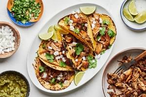 crispy carnitas tacos on a plate surrounded by bowls of chopped onion, cilantro, salsa verde and lime wedges for topping