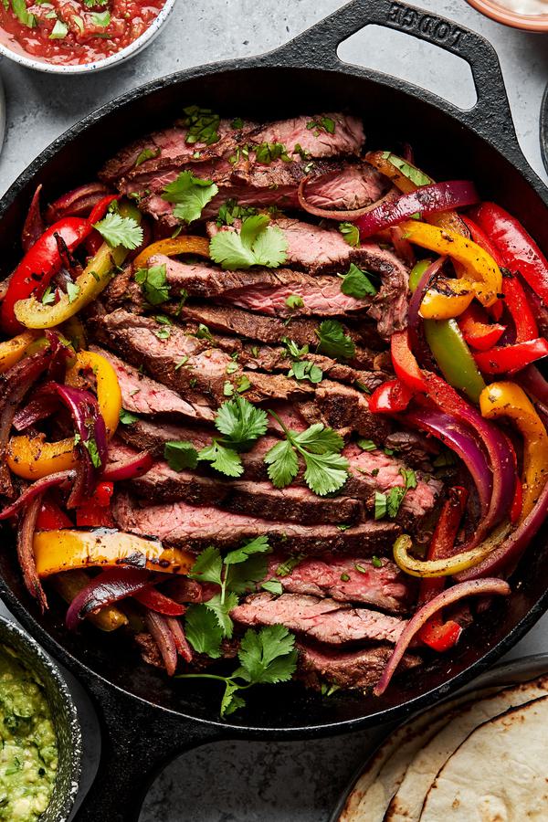 steak fajitas and peppers in a cast iron skillet sprinkled with cilantro surrounded by salsa, guacamole and corn tortillas